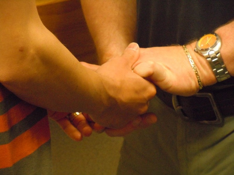 Close-up of two people's hands and forearms clasped together, dim warm lighting, one wrist wearing a watch and bracelet