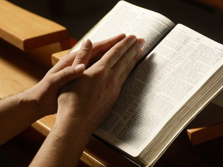 Close-up of hands clasped together resting on an open book on a wooden surface, warm golden lighting, reflective and peaceful