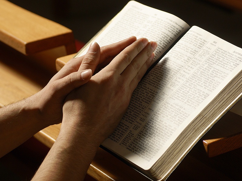 Close-up of hands clasped together on an open book resting on wooden pews, warm lighting, peaceful and reflective mood