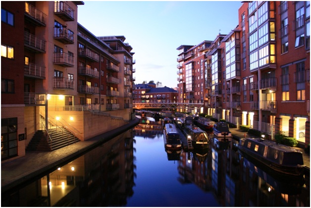 Nighttime cityscape of brick apartment buildings lining a canal with boats docked, lights reflecting on the water